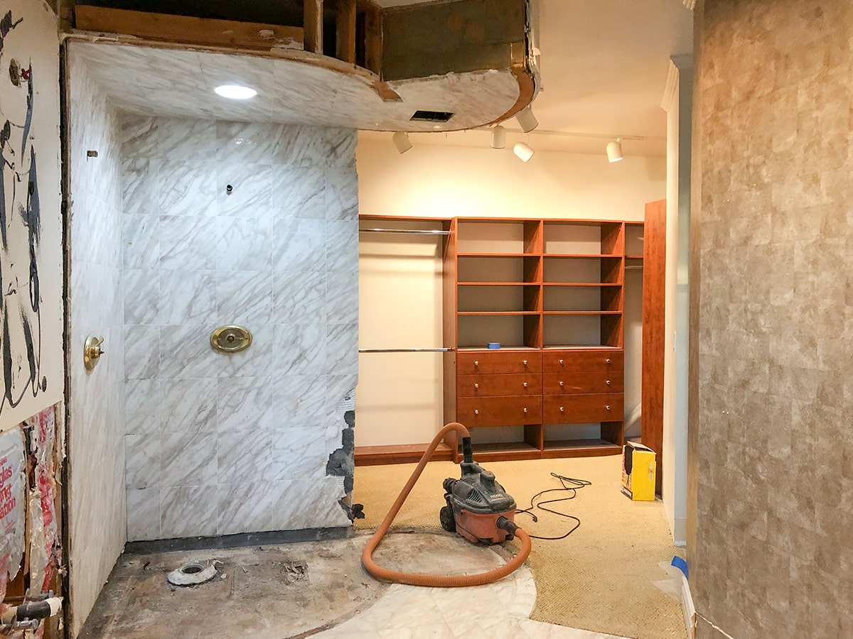 Interior room under renovation with missing flooring, marble wall, wooden shelves, and a wet/dry vacuum cleaner on the floor.