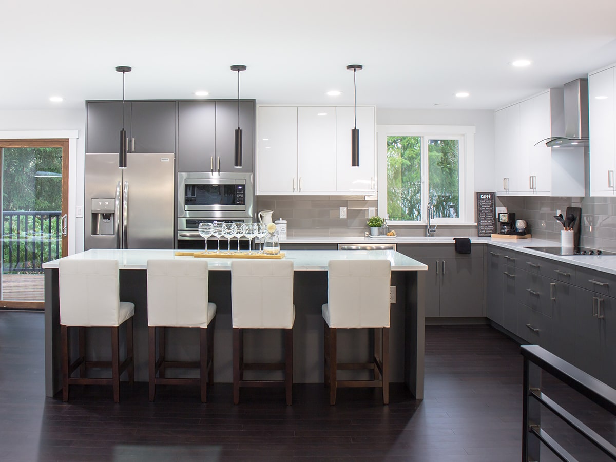 Modern kitchen with dark lower cabinets and white upper cabinets, a large island with four white cushioned stools, stainless steel refrigerator and appliances, pendant lights, and a window overlooking greenery.