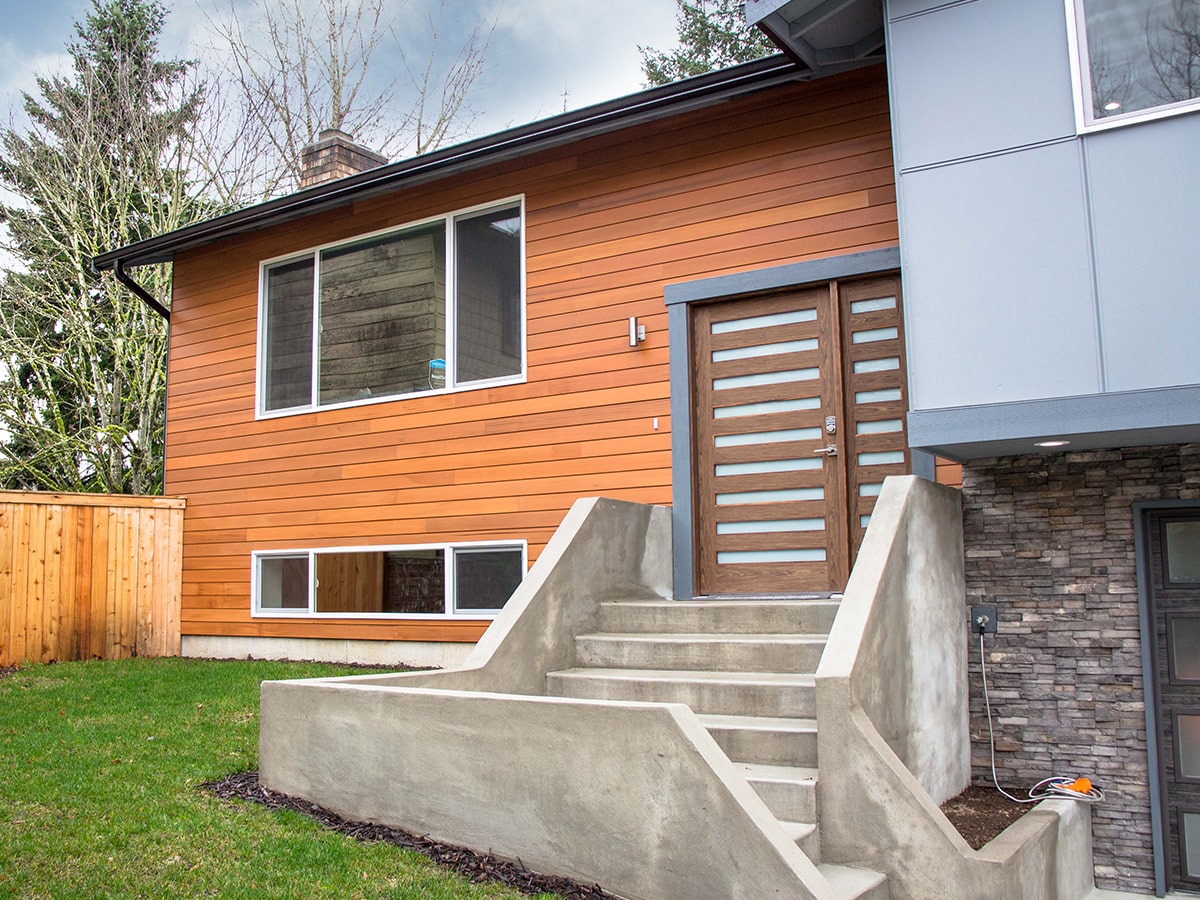 Modern house exterior with warm wood siding, concrete stairs leading to a wooden front door, green lawn, and stone accent wall.