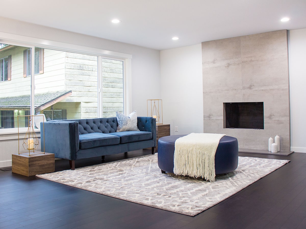 Modern living room with a blue velvet sofa, patterned rug, round navy ottoman with white throw, and a minimalist fireplace.