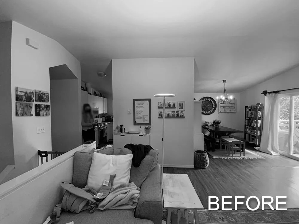 Black and white photo of a living and dining area with a sofa and pillows in the foreground, a standing lamp, and a dining table with chairs near sliding glass doors.