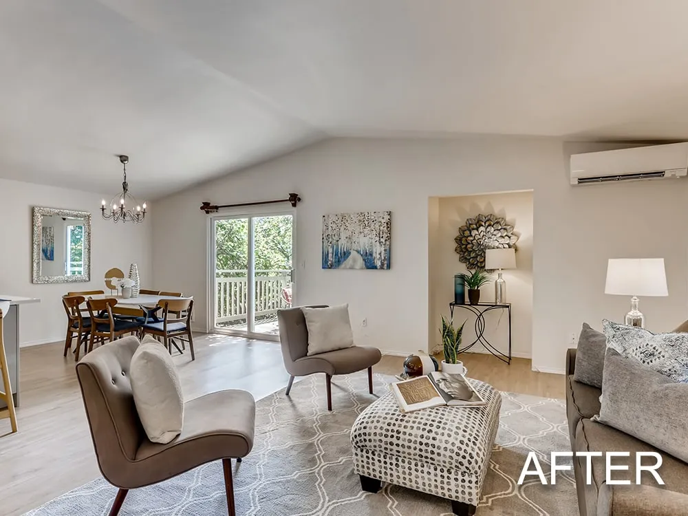 Modern open-plan living area with light wood floors, gray and beige furniture, a kitchen island with bar stools, stainless steel refrigerator, and large windows.