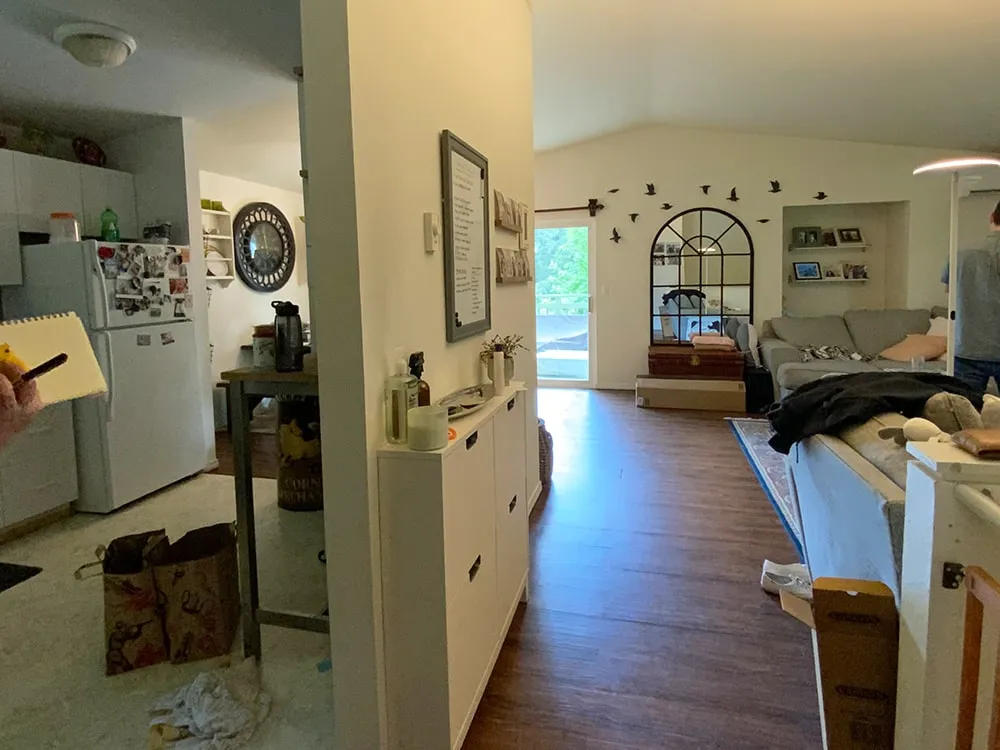 Interior view of a cozy living space with a kitchen on the left, wooden floors, a gray sofa, wall decor of birds, and a large window letting in natural light.