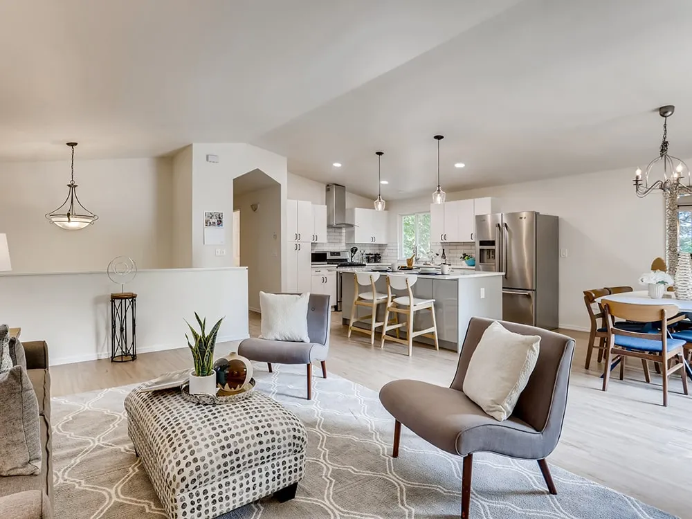 Modern open-concept living area with gray armchairs, patterned ottoman, and kitchen with white cabinets and stainless steel appliances.