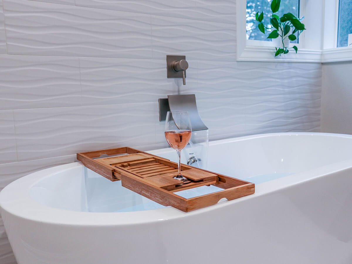 Close-up of relaxing bathtub setup with waterfall faucet and wooden bath caddy holding a glass of wine, styled for a luxurious bathroom retreat.