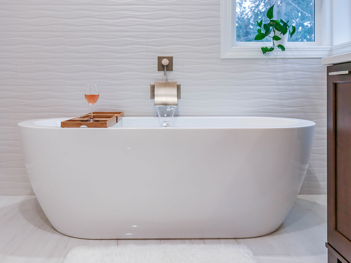 Freestanding bathtub with waterfall faucet and textured white tile wall in a bright, elegant bathroom remodel.