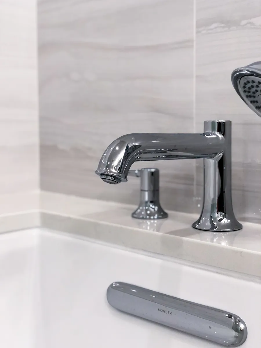 Close-up of a modern chrome bathtub faucet and handheld shower sprayer on a white quartz tub deck in a contemporary bathroom.