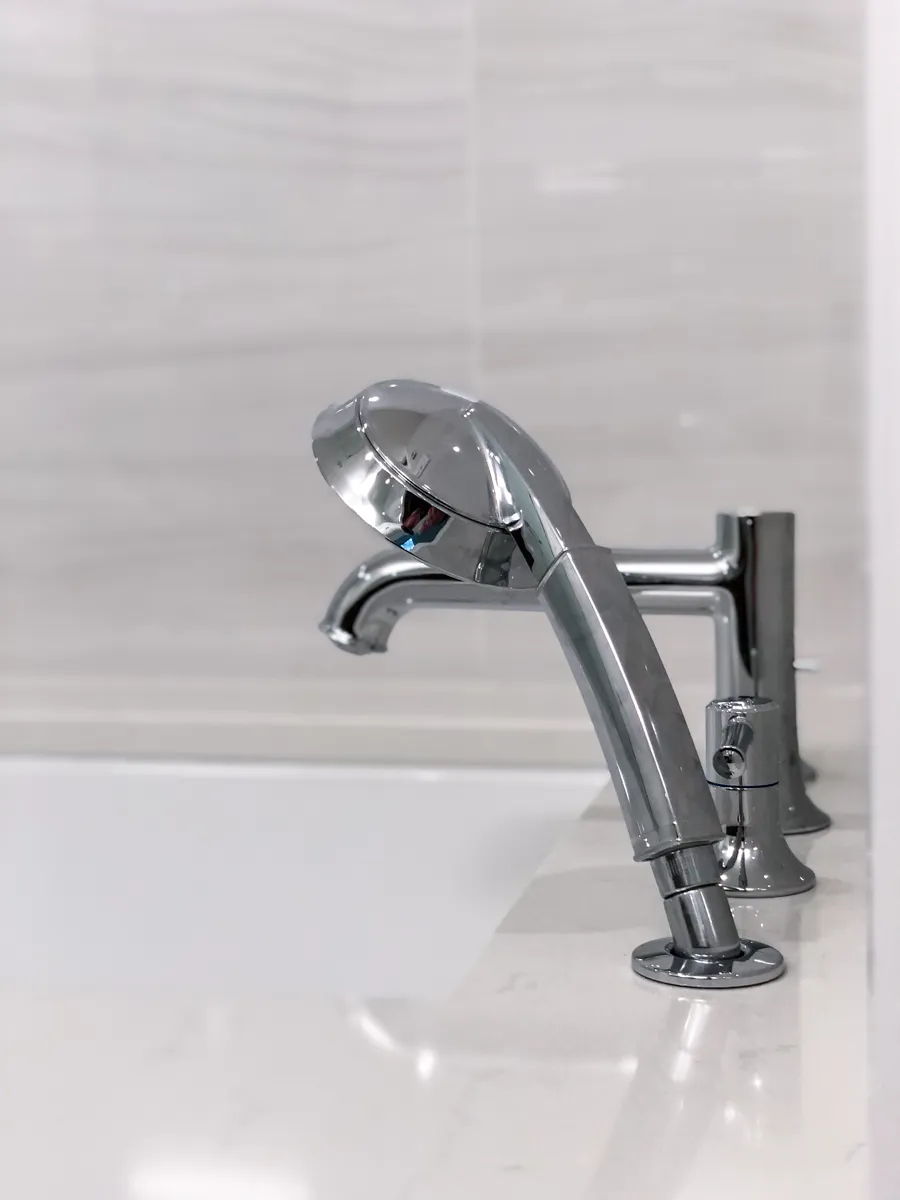 Close-up of a modern chrome bathtub faucet and handheld shower sprayer on a white quartz tub deck in a contemporary bathroom.