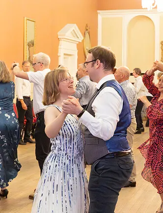 Couple smiling and dancing closely in a lively ballroom with other dancing guests.