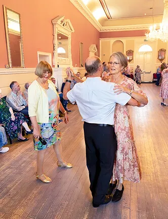 An elderly couple dancing in a large room while other seniors watch and smile.