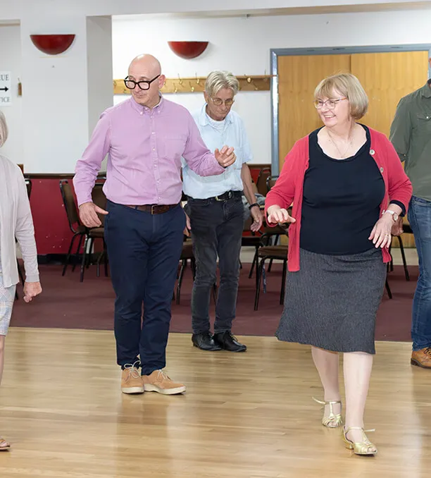 A group of older adults dancing in a community hall with wooden floor and chairs in the background.