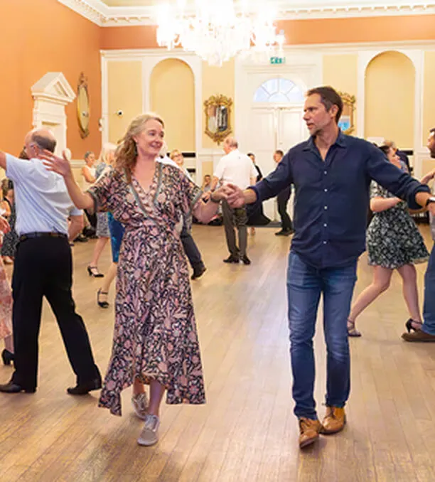 Several couples ballroom dancing in a brightly lit hall with wooden flooring and ornate walls.
