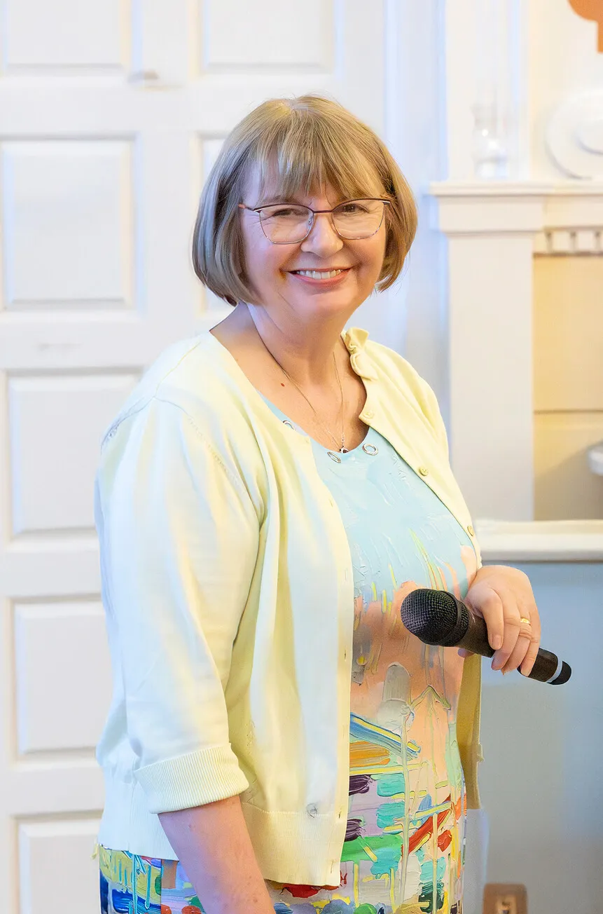 Smiling older woman wearing glasses, a light yellow cardigan, and a colorful dress holding a microphone indoors.