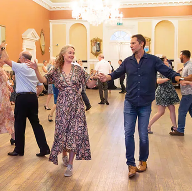 Couples dancing together in a spacious, well-lit ballroom with wooden floors and chandeliers.