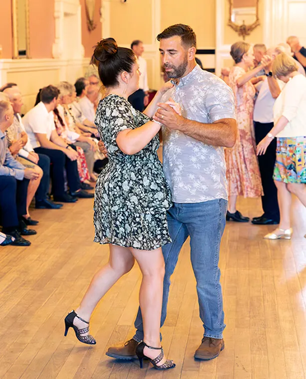 Couple dancing closely in a hall with an audience seated and more people dancing in the background.