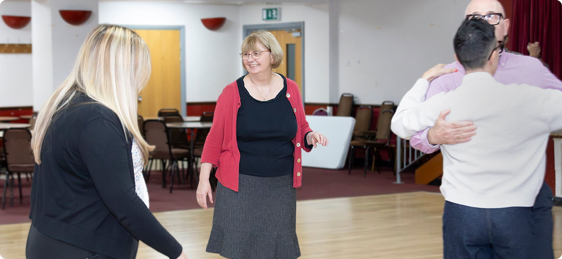 Four adults in a room with chairs and tables, three people are standing and one couple is dancing, smiling and interacting.