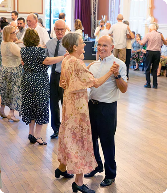 Elderly couples dancing in a spacious, well-lit room with wooden floors during a social event.