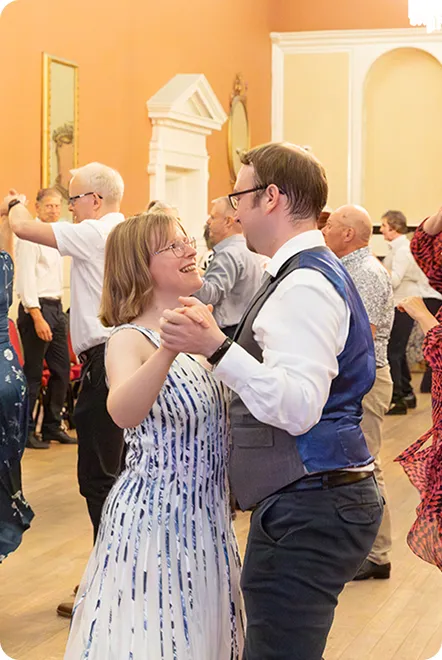 Couple smiling and dancing closely in a room with other dancers in the background.