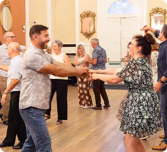 A group of people dancing together in a bright room with wooden floors and ornate mirrors on the walls.