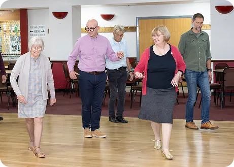 Five older adults walking and dancing together in a spacious room with wooden floor.