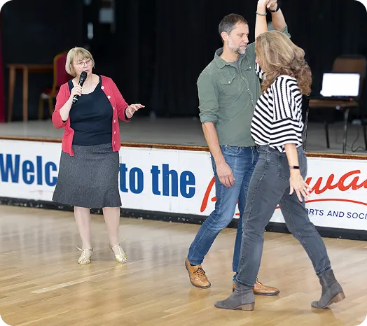 A woman holding a microphone speaking while a man in a green shirt and a woman in a striped top dance together on a wooden floor.