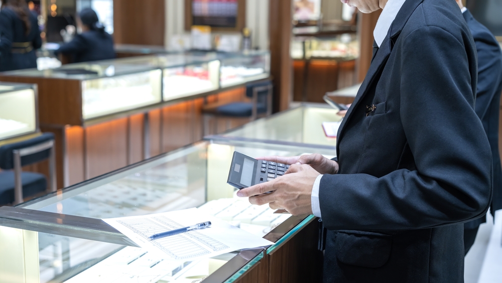 Male employees in suits using calculators and paperwork inside a jewelry store, performing inventory or financial tasks near display counters with luxury items.