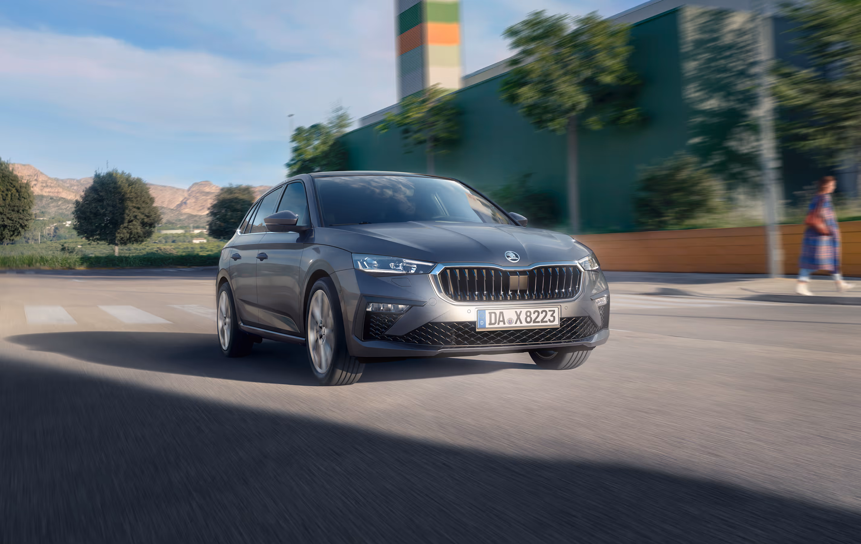 Eine dynamische Dreiviertelansicht von vorne.  Ein Škoda Scala in der Lackierung Graphite-Grau Metallic fährt auf einer Straße durch eine städtische Umgebung. Die Bewegungsunschärfe des Hintergrunds, in dem Bäume und Berge zu sehen sind, unterstreicht die Fahrszene.