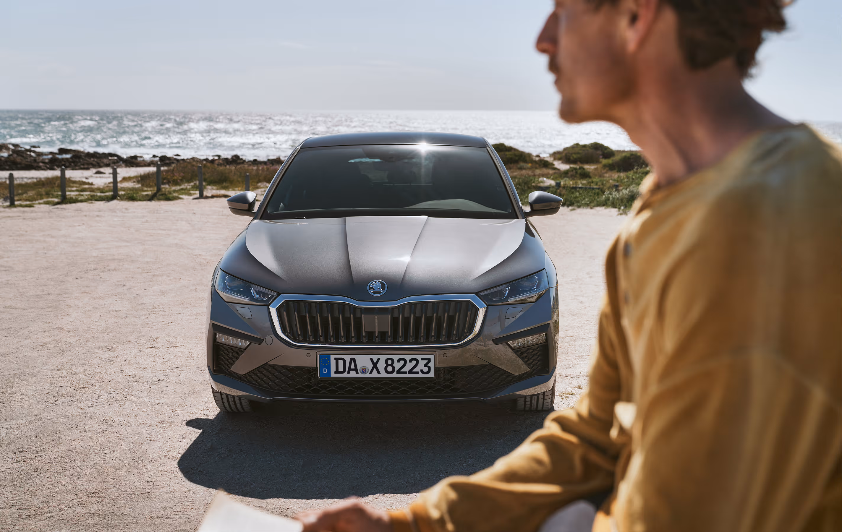 Ein Škoda Scala in der Lackierung Graphite-Grau Metallic ist frontal auf einem sandigen Parkplatz am Meer abgebildet. Im unscharfen Vordergrund steht eine Person, die auf das Fahrzeug blickt. Die Sonne spiegelt sich auf der Windschutzscheibe und dem Panoramaschiebedach.