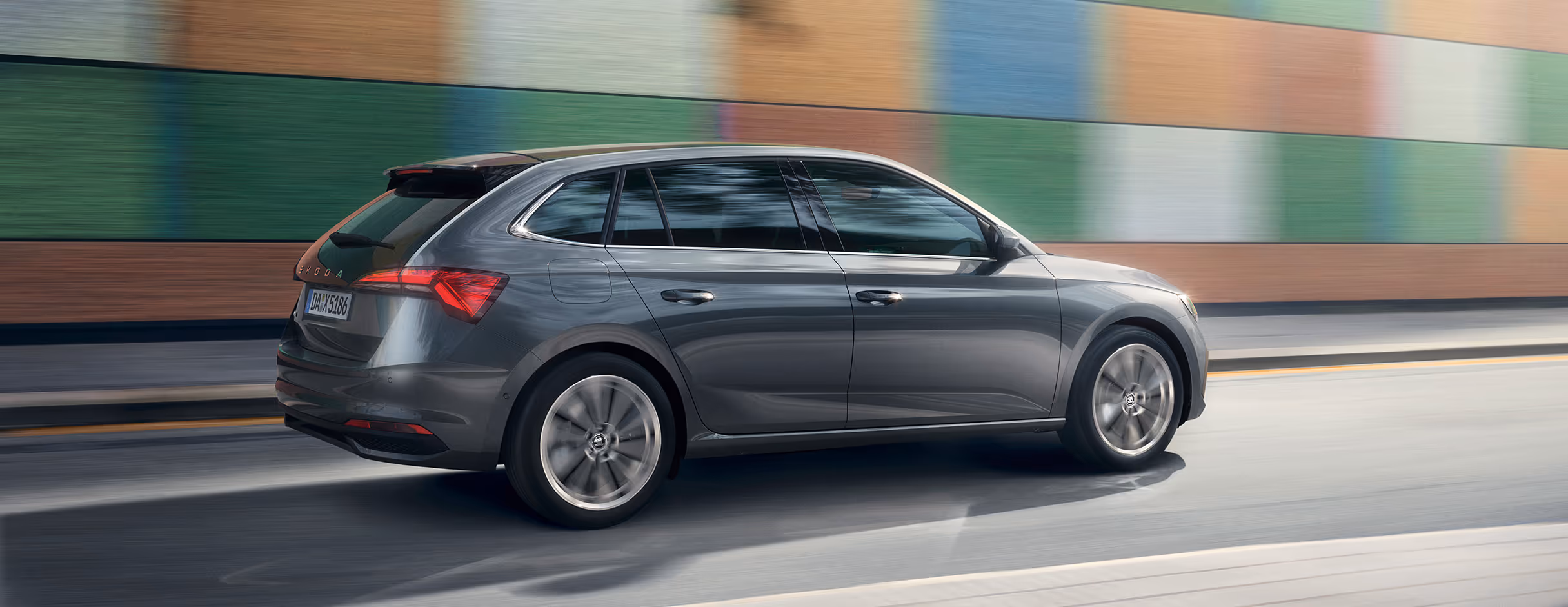 Ein Škoda Scala in der Lackierung Graphite-Grau Metallic fährt auf einer Straße vor einer bunt gemusterten Mauer entlang. Die Aufnahme zeigt das Fahrzeug in einer dynamischen Dreiviertelansicht von hinten. Die Bewegungsunschärfe unterstreicht die Fahrszene.