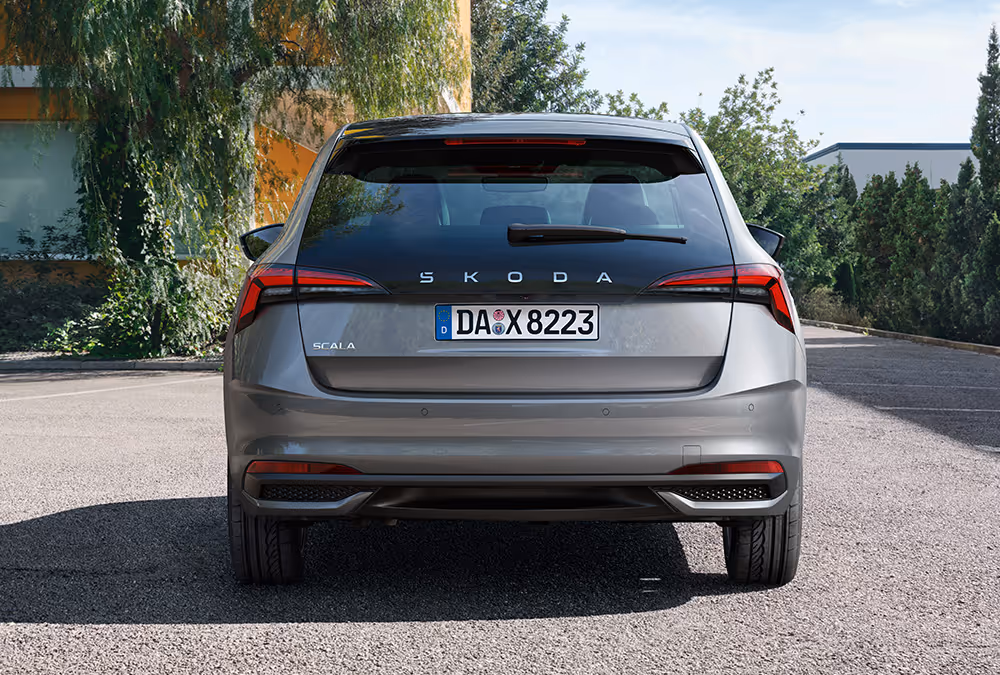 Eine frontale Heckansicht eines Škoda Scala in der Lackierung Graphite-Grau Metallic. Das Fahrzeug parkt auf einer asphaltierten Straße, im Hintergrund sind Bäume und ein Gebäude zu sehen.