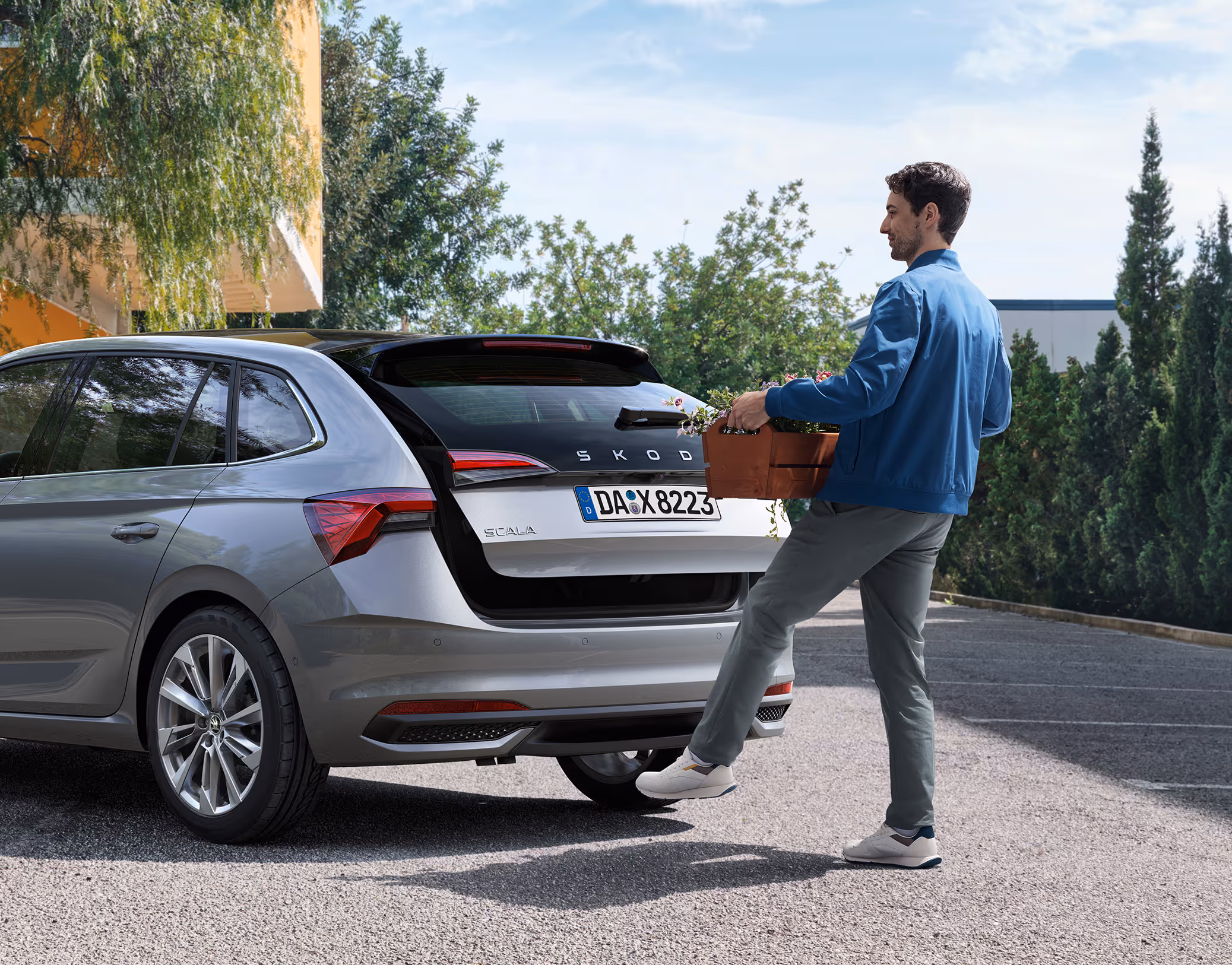 Ein Mann öffnet die elektrische Heckklappe eines Škoda Scala mit Hilfe des Virtuellen Pedals. Während er einen Blumenkasten in den Händen hält, löst er mit einer Fußbewegung unter dem Heckstoßfänger den Öffnungsmechanismus aus.