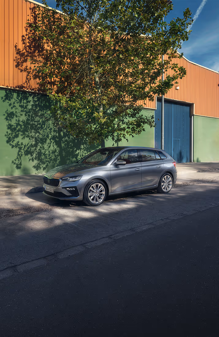 Vor einer grünen Wand mit Baumschatten parkt ein Škoda Scala Balance in der Lackierung Graphit-Grau Metallic. Die Schrägansicht von vorne präsentiert die sportliche Frontpartie und die eleganten Leichtmetallfelgen des Sondermodells. Das natürliche Lichtspiel auf der Karosserie unterstreicht das moderne Design des Kompaktwagens.