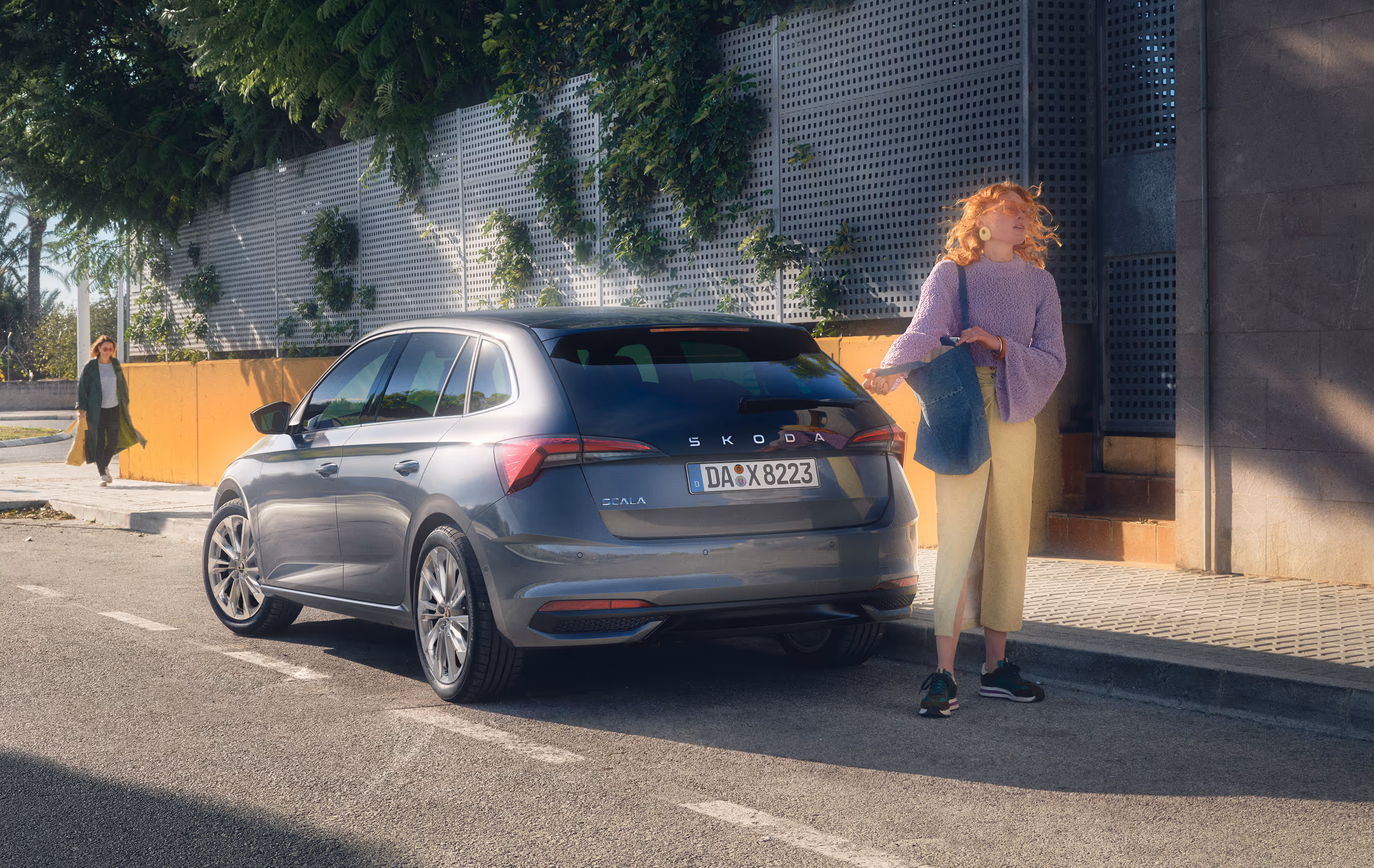 Ein Škoda Scala in der Lackierung Graphite-Grau Metallic ist am Straßenrand in einer sonnigen, städtischen Umgebung geparkt. Eine junge Frau steht am Heck des Fahrzeugs und scheint auf jemanden zu warten. Die Szene zeigt das Fahrzeug in einer Dreiviertelansicht von hinten, während im Hintergrund eine weitere Person auf dem Gehweg entlanggeht.
