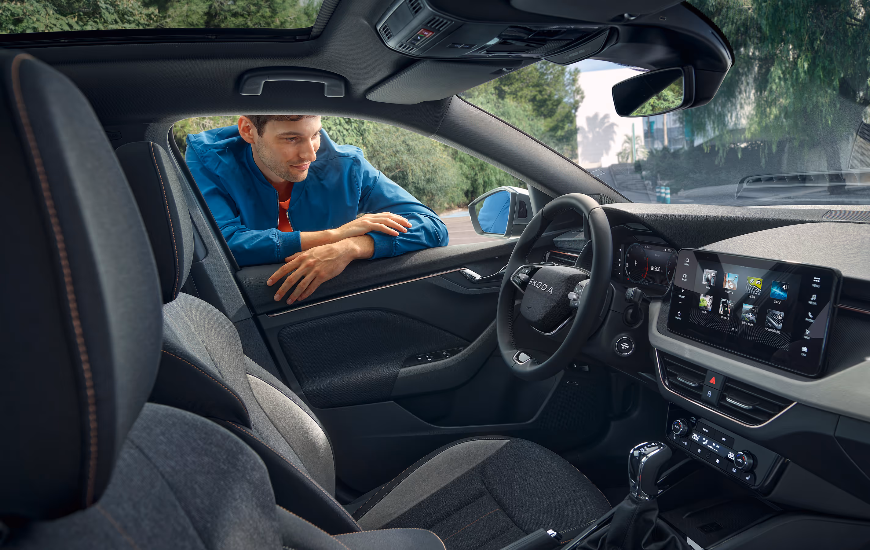 Ein Mann lehnt durch das geöffnete Fahrerfenster eines Škoda Scala und blickt in den Innenraum. Die Ansicht zeigt das Cockpit sowie die Vordersitze der Design Selection Lodge mit ihrer Stoff-Suedia-Kombination.