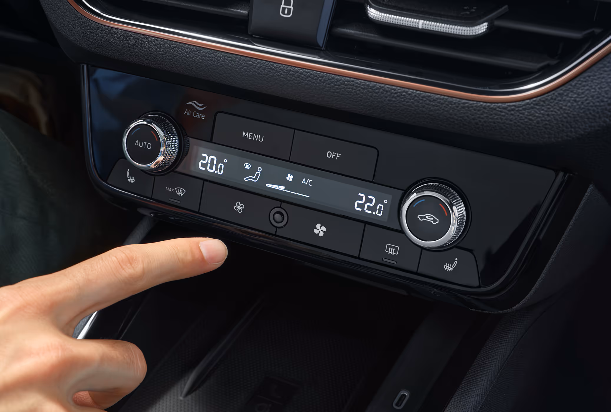 Eine Nahaufnahme zeigt das Bedienfeld der 2-Zonen-Klimaanlage Climatronic im Cockpit eines Škoda Scala. Ein Finger zeigt auf eine der Tasten unterhalb des zentralen Displays, auf dem die unterschiedlichen Temperatureinstellungen für Fahrer und Beifahrer angezeigt werden. Die Bedieneinheit ist Teil der Design Selection Lodge und wird von einer kupferfarbenen Dekorleiste akzentuiert.