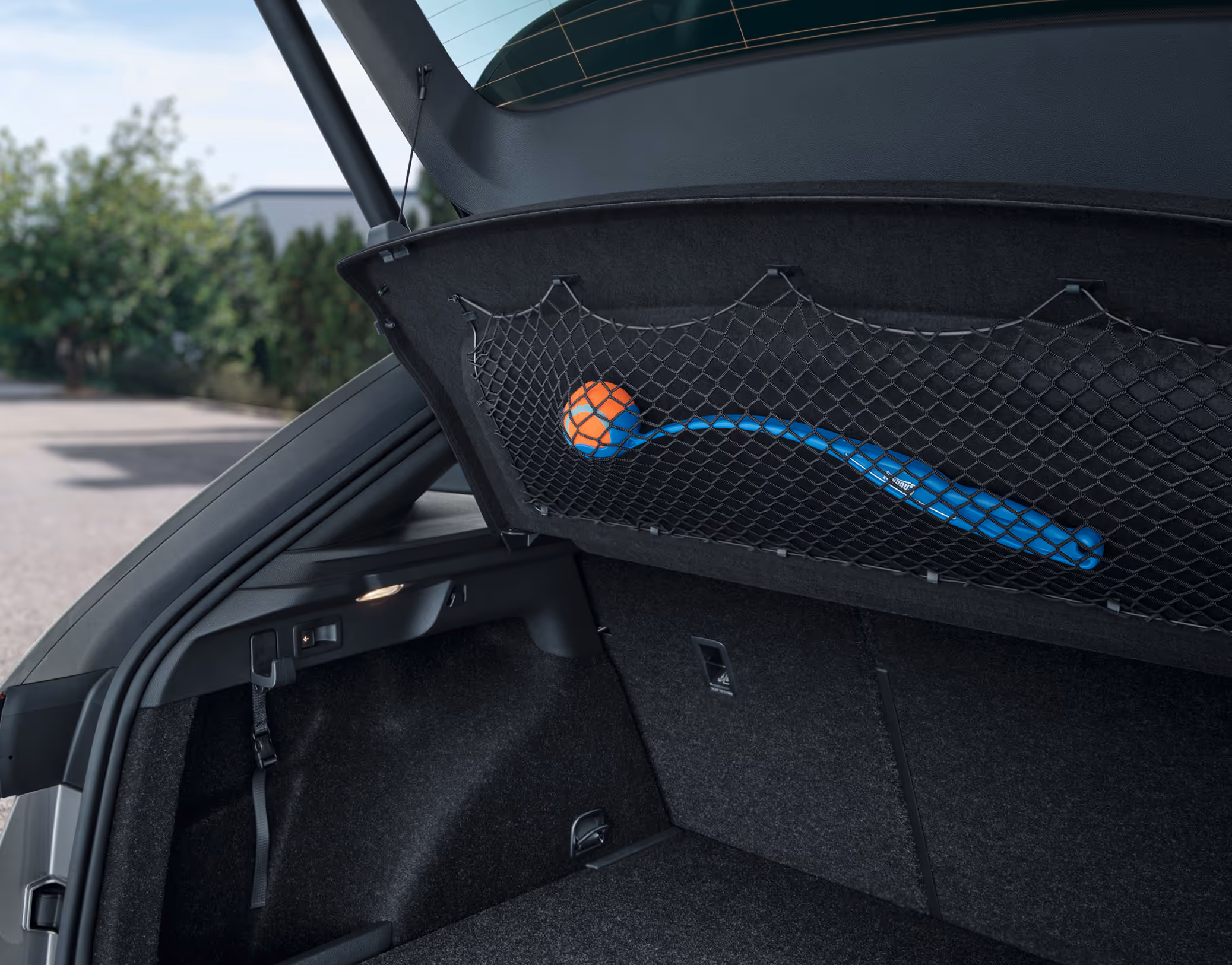 Eine Nahaufnahme zeigt die Unterseite der Gepäckraumabdeckung im Kofferraum eines Škoda Scala. An dieser Abdeckung ist ein schwarzes Gepäcknetz befestigt, das Teil des Netzprogramms ist. In dem Netz ist ein blaues Ballwurfspielzeug für Hunde verstaut.