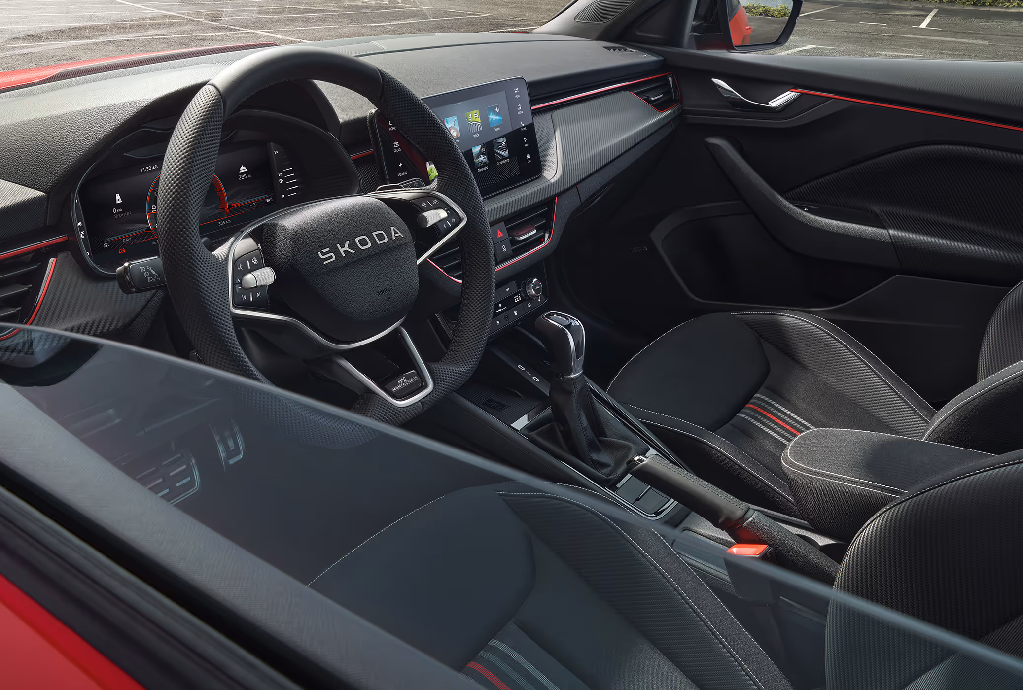 Ein Blick durch die geöffnete Fahrertür in das Cockpit eines Škoda Scala Monte Carlo. Das sportliche Interieur der Design Selection Monte Carlo zeigt schwarze Sportsitze mit roten und grauen Streifen, Dekorleisten in Carbon-Optik und eine rote Ambientebeleuchtung am Armaturenbrett.