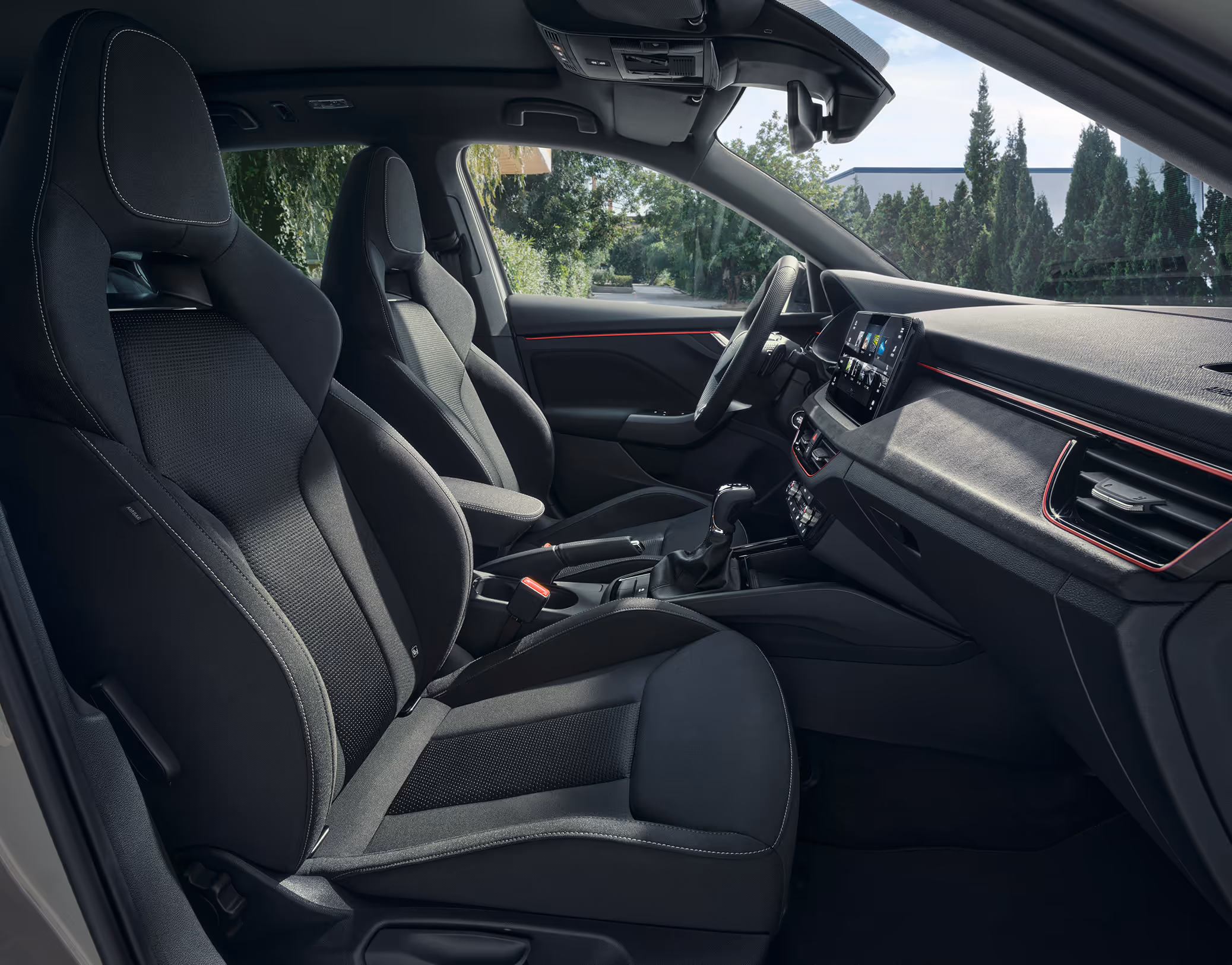 Eine seitliche Innenansicht des Škoda Scala zeigt die Vordersitze und das Cockpit mit der Design Selection Dynamic. Die Sportsitze mit integrierten Kopfstützen sind mit einer Kombination aus schwarzem Stoff und Microfaser-Stoff (Suedia) bezogen und werden durch rote Akzente im Interieur sowie weiße Kontrastnähte ergänzt.