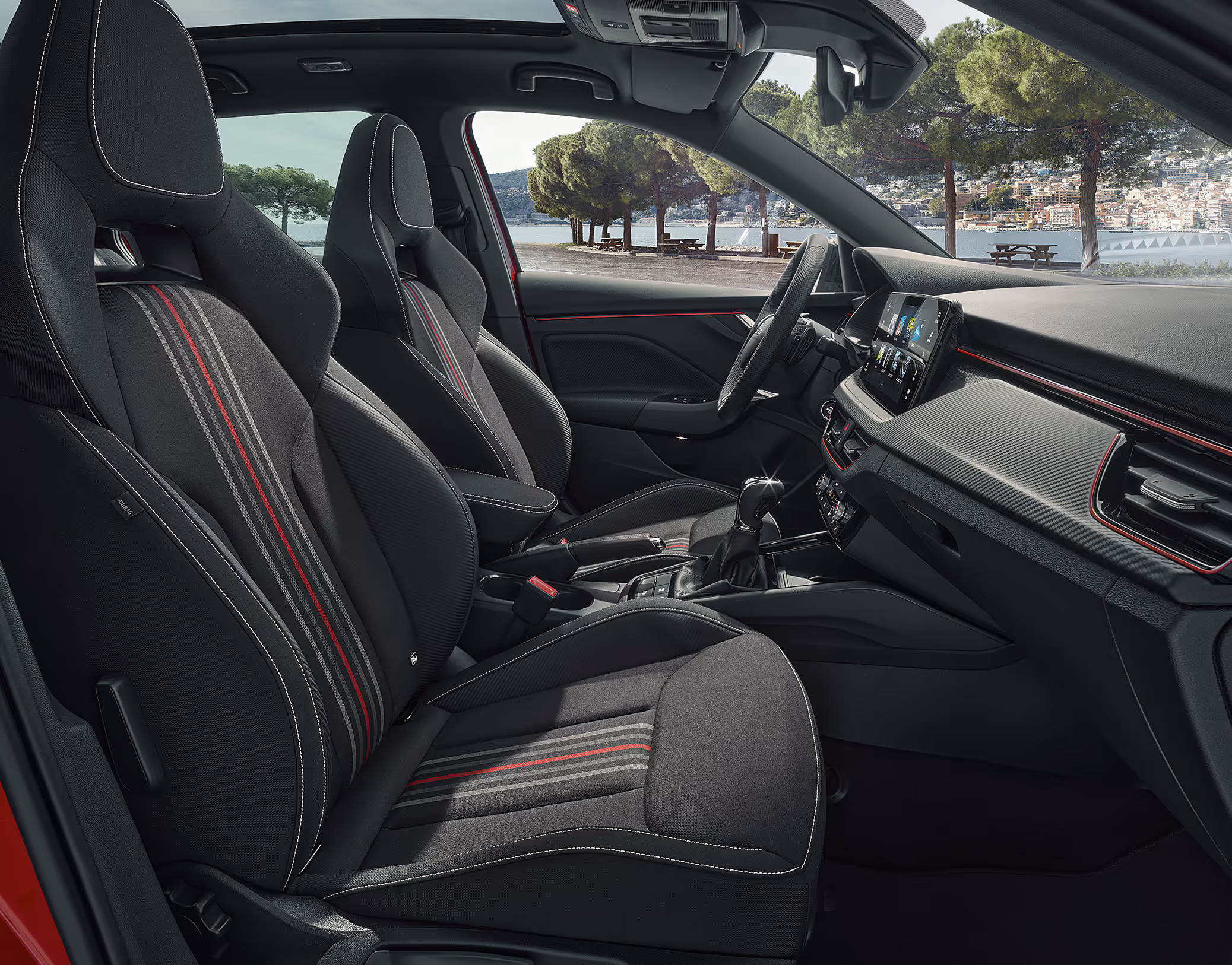 Eine seitliche Innenansicht des Škoda Scala Monte Carlo zeigt das sportliche Interieur der Design Selection Monte Carlo. Die schwarzen Sportsitze mit integrierten Kopfstützen sind mit roten und grauen Zierstreifen versehen und werden durch eine rote Ambientebeleuchtung und Dekorleisten in Carbon-Optik ergänzt. Durch die Fenster ist eine Küstenlandschaft zu sehen.