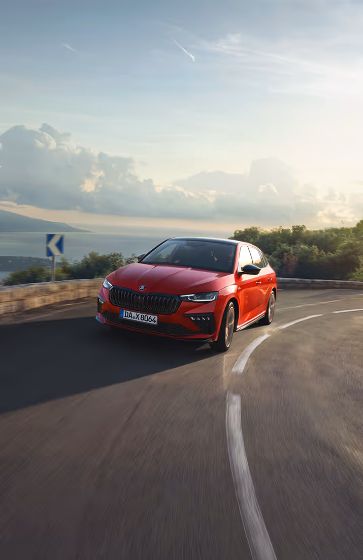 Ein Škoda Scala Monte Carlo in der Lackierung Velvet-Rot Metallic fährt auf einer kurvenreichen Küstenstraße in der Abendsonne. Das Fahrzeug wird in einer dynamischen Dreiviertelansicht von vorne gezeigt, während es eine Kurve durchfährt. Im Hintergrund sind das Meer und eine Küstenlinie zu erkennen.
