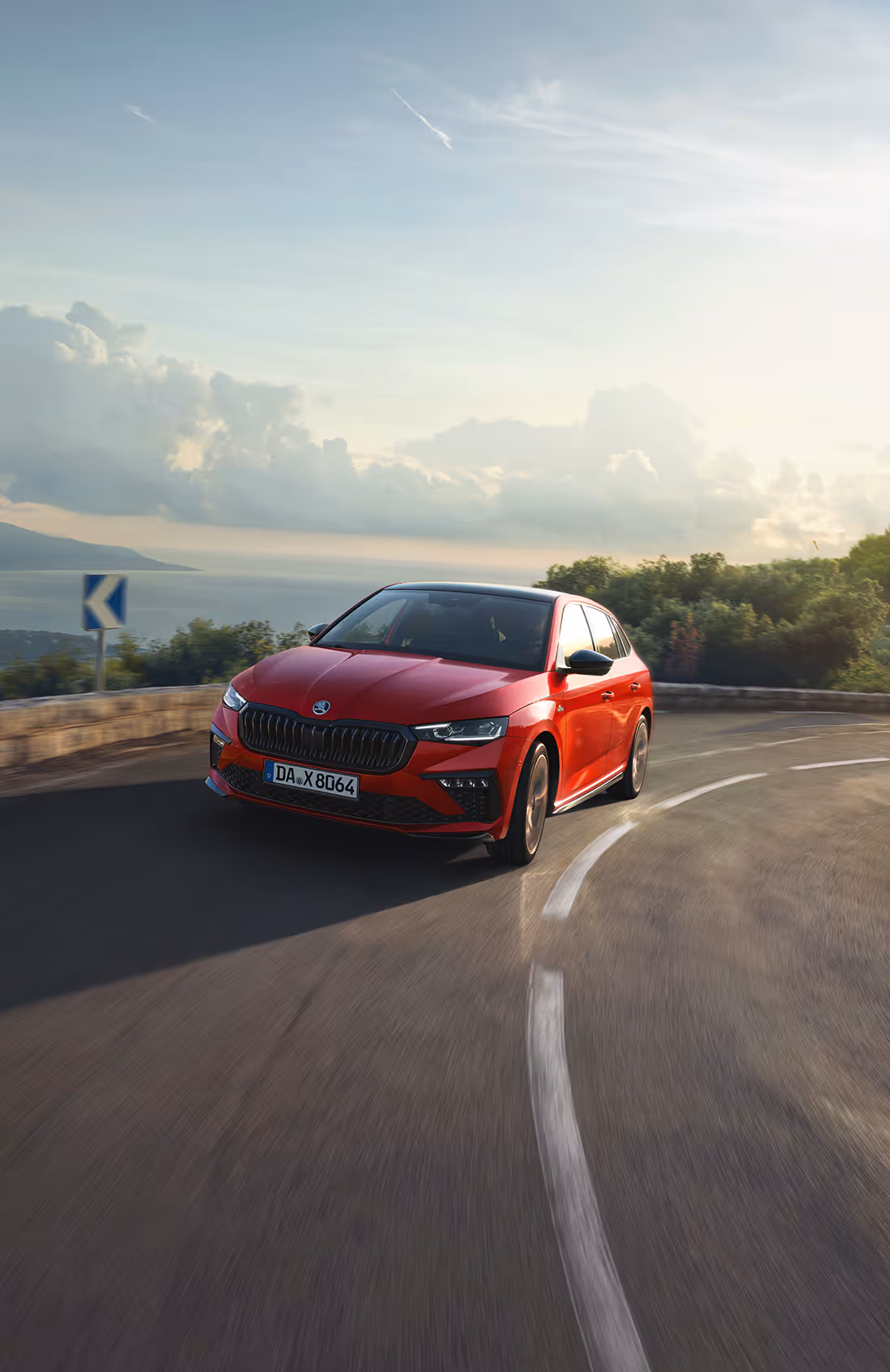 Ein Škoda Scala Monte Carlo in der Lackierung Velvet-Rot Metallic fährt auf einer kurvenreichen Küstenstraße in der Abendsonne. Das Fahrzeug wird in einer dynamischen Dreiviertelansicht von vorne gezeigt, während es eine Kurve durchfährt. Im Hintergrund sind das Meer und eine Küstenlinie zu erkennen.