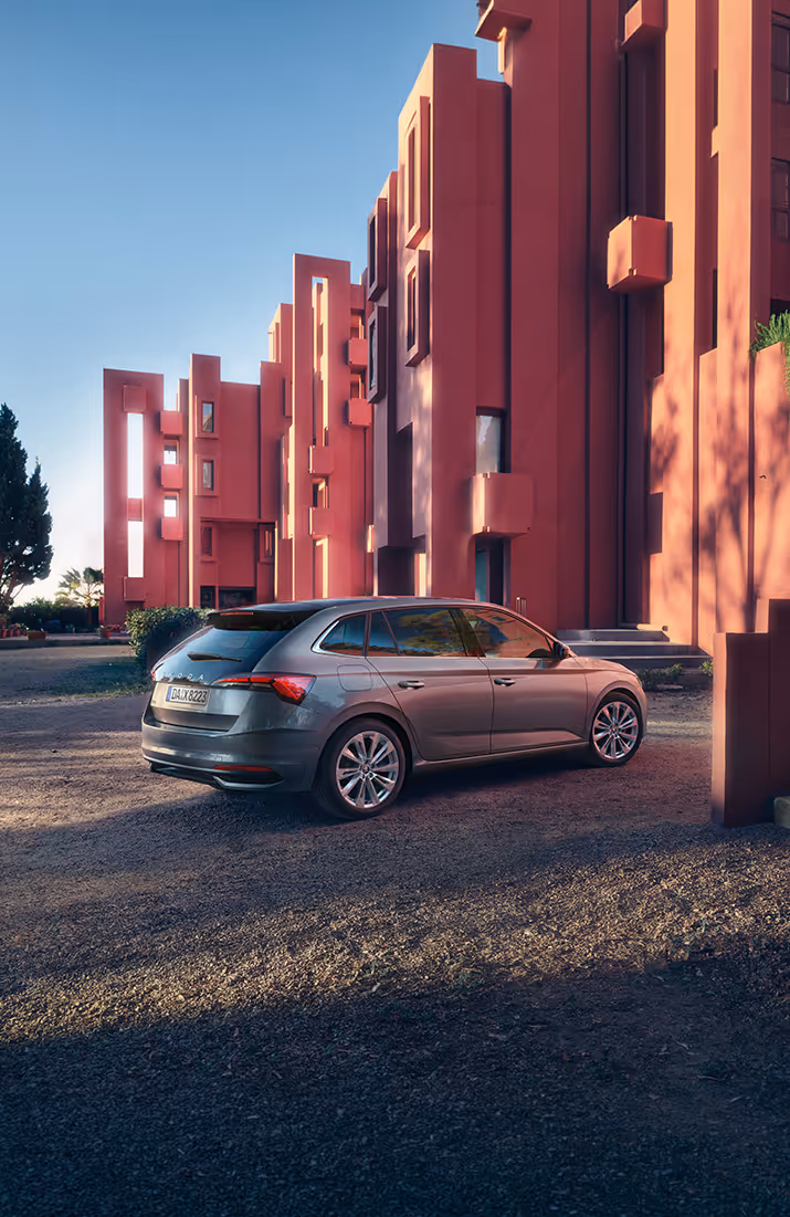 Schräge Heck-Seitenansicht auf den Škoda Scala Balance in der Lackierung Graphit-Grau Metallic vor einer architektonischen Kulisse in Terrakotta-Tönen. Markante Merkmale sind die verlängerte Heckscheibe, die abgedunkelten Scheiben im Fond sowie die hochwertigen Leichtmetallfelgen. Das Lichtspiel auf der Flanke unterstreicht die Linienführung des kompakten Fahrzeugmodells.