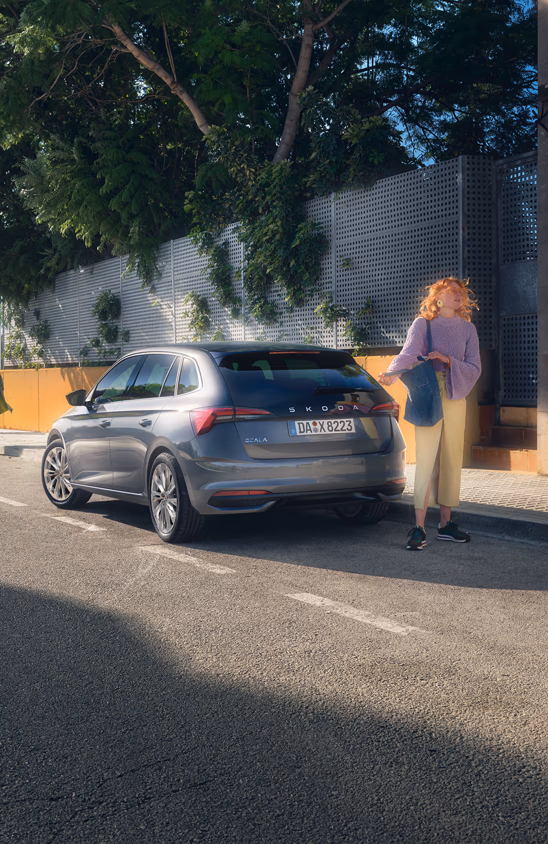 Ein Škoda Scala in der Lackierung Graphite-Grau Metallic ist am Straßenrand in einer sonnigen, städtischen Umgebung geparkt. Eine junge Frau steht am Heck des Fahrzeugs und scheint auf jemanden zu warten. Die Szene zeigt das Fahrzeug in einer Dreiviertelansicht von hinten, während im Hintergrund eine weitere Person auf dem Gehweg entlanggeht.