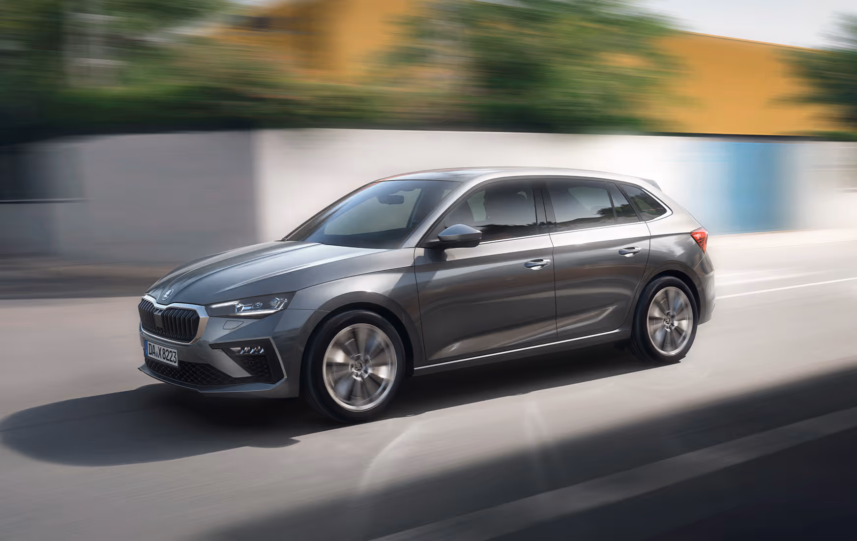 Dynamische Fahr-Aufnahme eines Škoda Scala in der Lackierung Graphite-Grau Metallic. Das Bild zeigt die Front-Schrägansicht des Kompaktwagens in Bewegung auf einer städtischen Straße. Durch den Mitzieher-Effekt verschwimmt der Hintergrund und betont die Dynamik des Fahrzeugs.