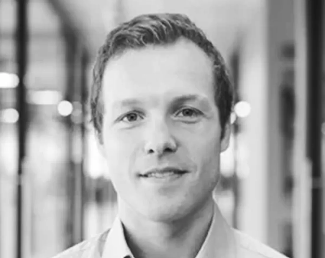 Close-up black and white photo of a smiling man with short hair wearing a collared shirt in an indoor setting.