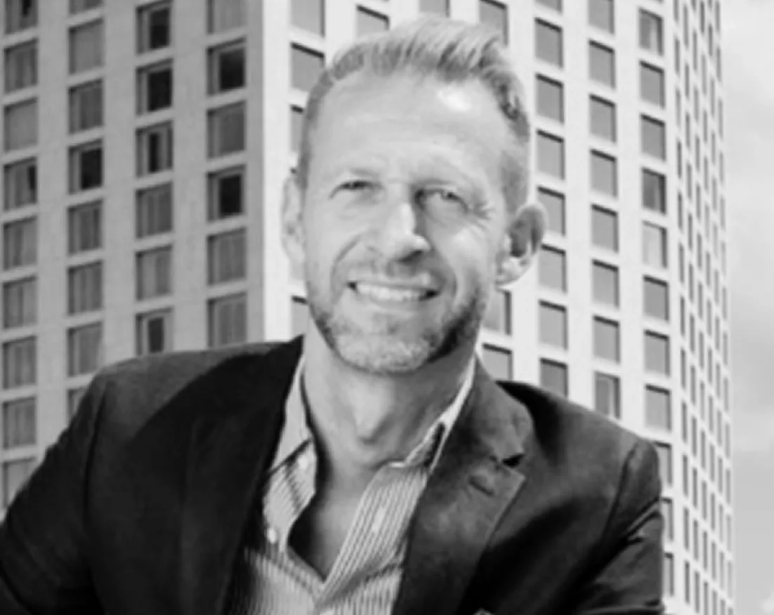 Smiling man in a suit jacket and striped shirt posing outdoors in front of a modern building.