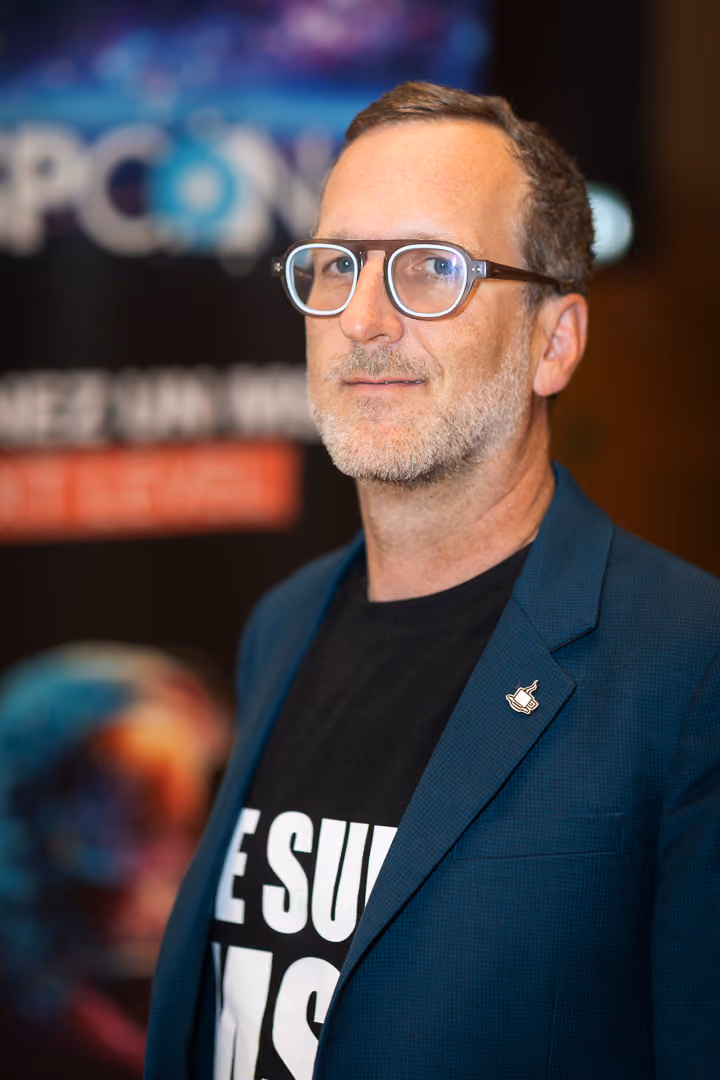 Homme portant des lunettes à monture épaisse, un blazer bleu et un t-shirt noir avec un texte partiellement visible, sur fond flou avec des lettres et une image colorée.