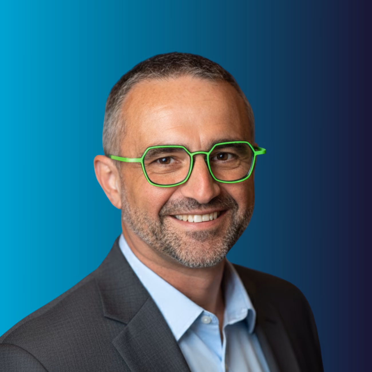 Homme souriant portant un costume gris, une chemise bleue claire et des lunettes vertes originales.