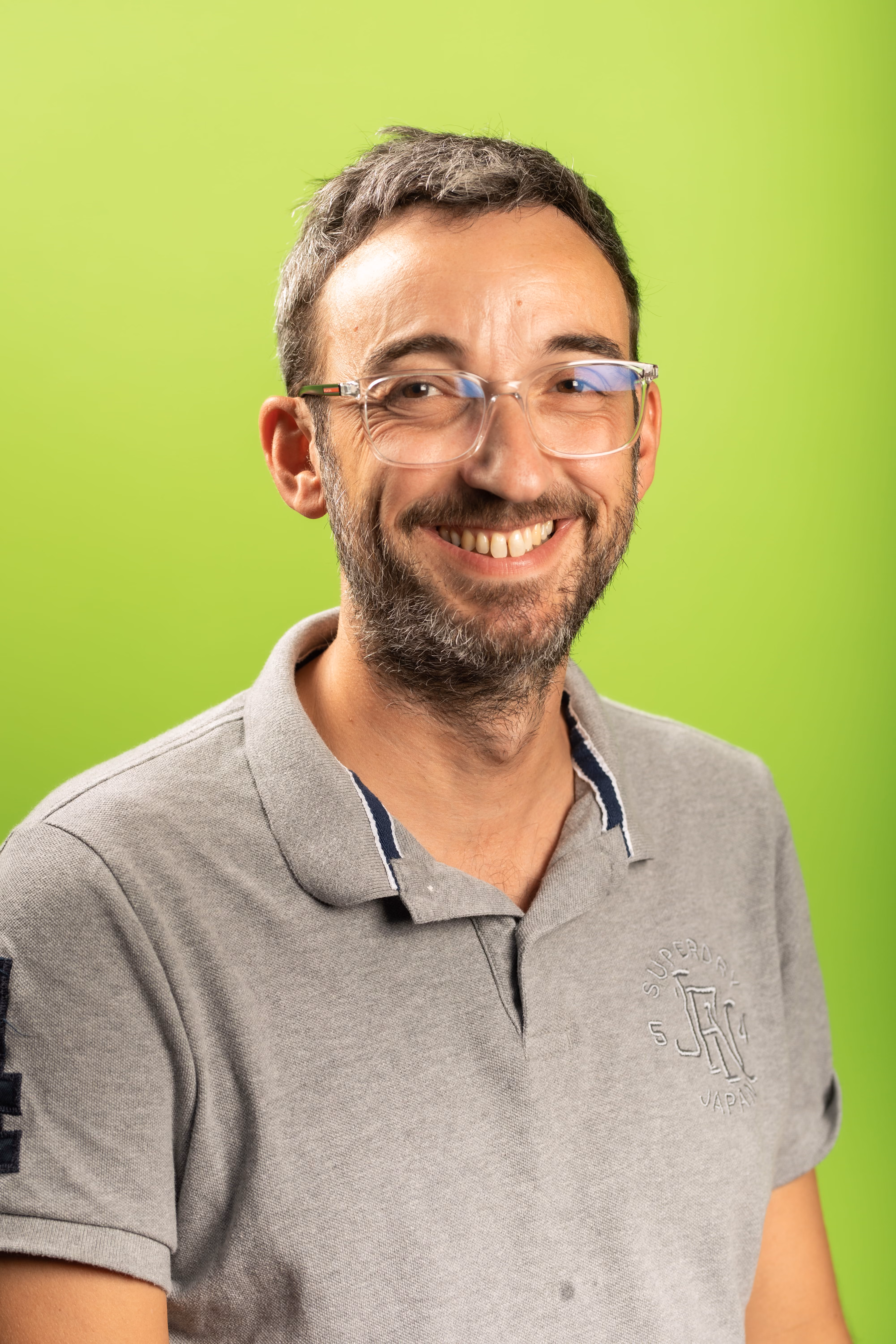 Homme souriant portant des lunettes transparentes et un polo gris devant un fond vert clair.