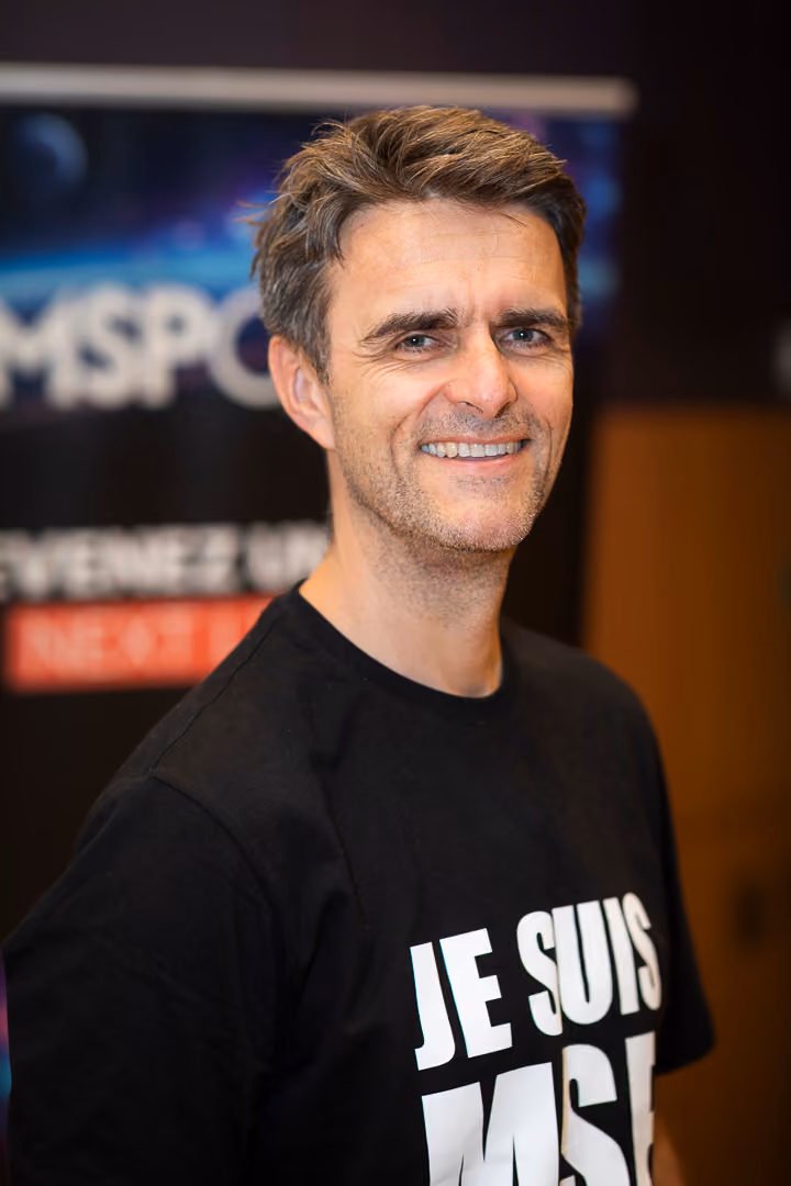 Homme souriant portant un t-shirt noir avec l'inscription partielle « JE SUIS » en lettres blanches.